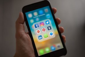 A man holds his Iphone and shows social media channels that he uses daily: Facebook, Instagram, Twitter, Pinterest, LinkedIn, Whatsapp, Messenger, tumblr and YouTube.